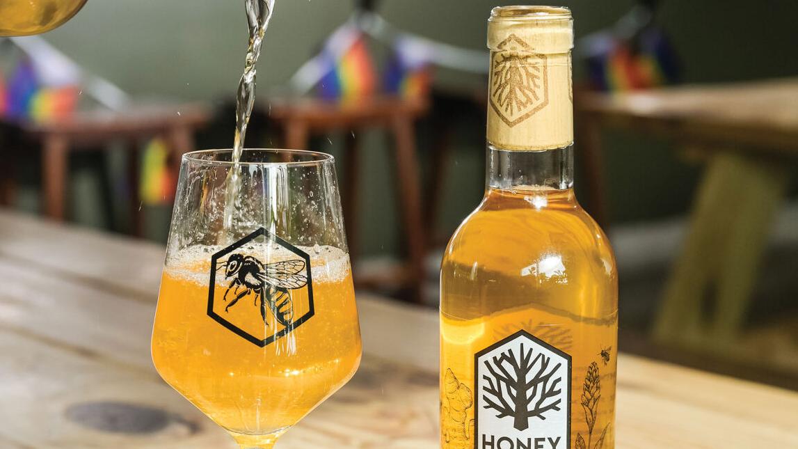 A bottle of Sweet Baby Ginger from Honeytree Meadery placed next to a glass that is being filled by another bottle poured from above it.
